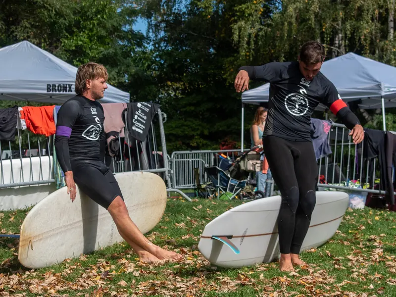 Deux surfeurs en combinaison marchent vers la rivière, planches sous le bras, devant le public